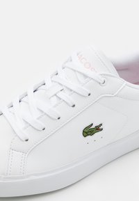 Sneaker bianco con finitura in pelle liscia, caratterizzato da un logo di coccodrillo ricamato in verde, lacci bianchi piatti e una suola in gomma testurizzata.