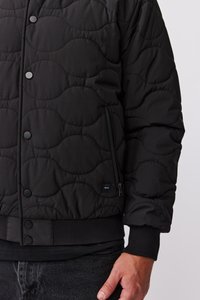 Veste en quilt noir avec un motif en vagues, dotée de boutons-pression, d'une poche zippée et de poignets et ourlet côtelés. Le tissu semble lisse et doux.