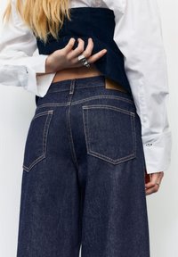 Oseba, oblečena v temno modre široke kavbojke in belo srajco, dviguje mornarsko moder telovnik, da razkrije trebuh in pokaže srebrne prstane na prstih.