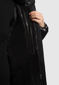 Melns pūšamais mētelis ar spīdīgu, quiltu iekšpusi. Iezīmējas ar ribotu melnu audumu pie apkakles un ar rāvējslēdzēju priekšpusē.