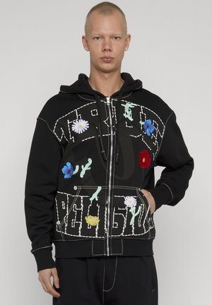 Giovane che indossa una felpa con cappuccio nera con cerniera, cuciture bianche e ricami floreali colorati, in piedi davanti a uno sfondo semplice.