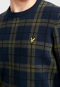 Pull en tricot avec un motif carreaux bleu marine et vert olive, doté d'un col côtelé et d'un petit emblème d'oiseau jaune sur la poitrine.