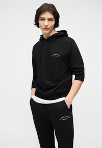 Νεαρός ενήλικας με μεσαίου μήκους σκουρόχρωμα μαλλιά, φορώντας μαύρη μπλούζα με κουκούλα και μαύρο παντελόνι φόρμας με λευκά λογότυπα, με τα χέρια στις τσέπες.