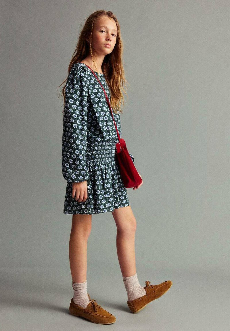 Deklica z dolgimi lasmi, oblečena v temno cvetlično obleko, v bež nogavicah, rjavih čevljih in nosi rdečo torbico čez ramo, na enobarvni sivi podlagi.