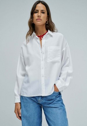 Frau trägt ein locker sitzendes weißes Hemd mit Brusttasche, blaue Jeans, eine rote Halskette und goldene Creolen, steht mit einer Hand in der Tasche.