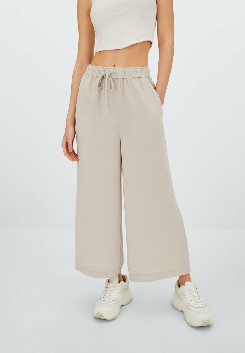 Femme portant un pantalon beige à jambes larges avec cordon de serrage, un crop top blanc et des baskets blanches épaisses, debout avec une main dans la poche.