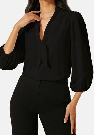 Femme portant un chemisier noir à manches longues avec un détail noué au col et un pantalon noir assorti, posant avec une main près de la clavicule et l'autre sur le côté.