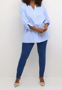 Frau trägt eine hellblaue Langarmbluse, dunkelblaue Skinny Jeans und beiges, spitzes Ballerina-Schuhwerk mit Schleifchen, während sie vor einer weißen Wand steht.