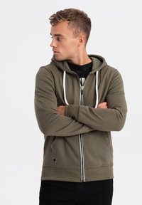 Olivově zelená mikina na zip s kapucí se stahovací šňůrkou, předními kapsami, bílými akcenty a žebrovanými manžetami. Model má ruce zkřížené a pod ní nosí černé tričko.