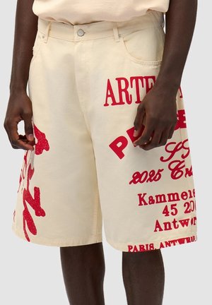 Shorts beige arrivant au genou en coton, ornés de textes rouges vifs et de motifs graphiques, avec des poches et une fermeture à bouton à la taille.