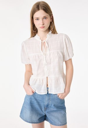 Jeune femme aux longs cheveux blonds porte un chemisier blanc texturé avec des manches bouffantes et un short en jean bleu, mains dans les poches.