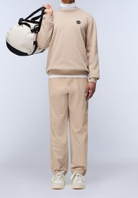 Hombre con suéter de cuello alto beige y pantalones, sosteniendo un bolso de deporte beige y negro sobre el hombro, con zapatillas blancas, sobre un fondo liso.
