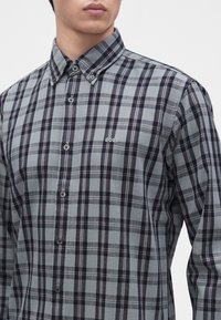 Homme portant une chemise à carreaux grise et noire boutonnée avec un col boutonné et le logo "BOSS" sur la poitrine.