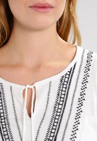 Weiße Bluse mit aufwendiger schwarzer Stickerei entlang der Vorderseite, mit rundem Ausschnitt und Bindeband am Hals.
