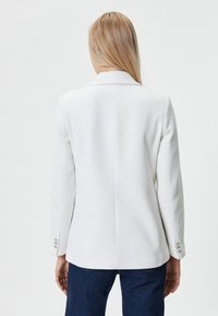 Blazer bianco a trama con colletto a rever e polsini a un bottone, caratterizzato da una vestibilità sartoriale e una finitura liscia all'esterno.