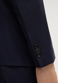 Manica di un blazer navy in tessuto testurizzato, con quattro bottoni neri al polsino, caratterizzata da filo abbinato e un taglio pulito e sartoriale.
