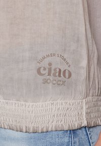 Helles beiges Oberteil mit gerafftem Saum, bedrucktem Text "SUMMER STORIES ciao SOCCX." Weiches Material mit subtiler Textur.