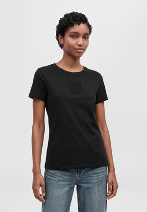 Un modèle aux cheveux courts et bouclés porte un tee-shirt noir à col rond avec un écusson carré sur la poitrine et un jean bleu, sur un fond uni et clair.
