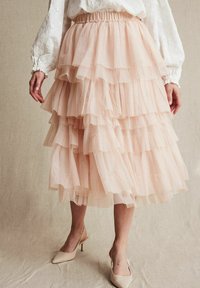 Lyserød tylskørt med lagdelte flæser, elastisk talje og en flydende design. Hvid bluse med blomsterbroderi synligt øverst.