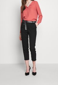 Kvinna iförd en korallfärgad långärmad blus, svarta cropped byxor med metallicbälte och svarta spetsiga klackar, stående mot en neutral bakgrund.