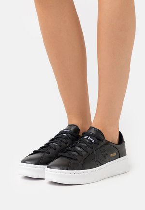 Sneakers in pelle nera con suola bianca, dotati di lacci neri, accenti perforati e dettaglio del logo dorato. Design classico a taglio basso.