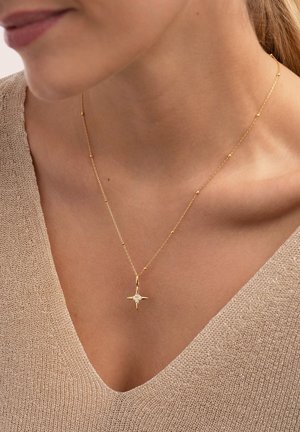 Collar de oro con un colgante en forma de estrella con una piedra central, cadena fina con pequeñas cuentas, combinado con una blusa beige texturizada.