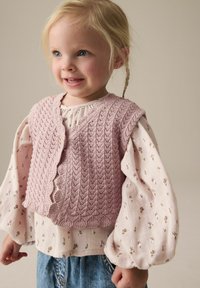 Gebreid lichtroze vest met geribde randen over een bloemetjes patroon blouse met lange mouwen. De blouse heeft pofmouwen en is gecombineerd met denim jeans.