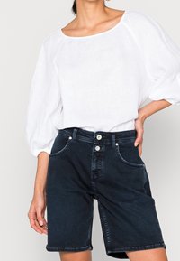 Shorts en denim bleu foncé à coupe classique, avec deux poches avant et une taille à double bouton. Associés à un chemisier blanc texturé.