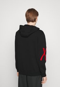 Schwarzer Kapuzenpullover mit langen Ärmeln, der ein großes rotes "X"-Akzent auf dem linken Arm hat. Hergestellt aus weichem Stoff mit einer strukturierten Oberfläche.