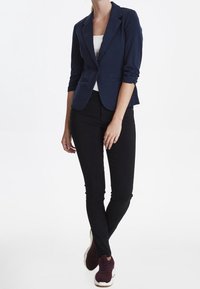 Mörkblå blazer med en figurnära design, som matchas med svarta slim-fit byxor och vinröda sneakers. Blazern har uppvikt ärmar och detaljer på slaget.