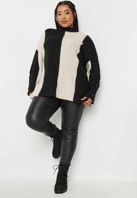 Czarny i kremowy, oversize sweter z pionowymi paskami, prążkowaną teksturą, wysokim kołnierzem oraz detalami w postaci guzików po bokach, zestawiony z czarnymi skórzanymi spodniami.