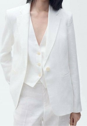Person iført en hvid skræddersyet blazer, matchende vest med tre knapper og hvide bukser mod en ensfarvet lys baggrund.
