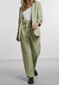 Jasnozielona marynarka i dopasowane spodnie z paskiem do wiązania, o gładkiej fakturze. Zestaw zawiera biały dopasowany top i czarne buty.