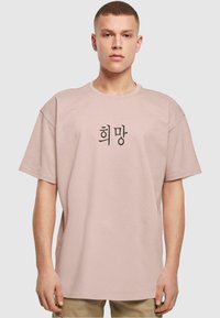 Korte mouwen, pastelroze t-shirt van katoen, met een zwarte Koreaanse karaktersprint in het midden en een losse, oversized pasvorm.