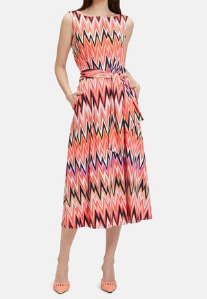 Robe midi sans manches avec un motif zigzag rose, orange, noir et blanc, ceinturée à la taille, avec des poches latérales, portée avec des escarpins corail à bout pointu.