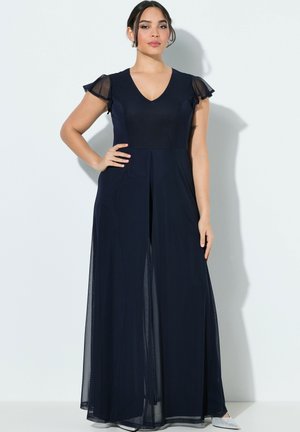 Kobieta w granatowej, sięgającej podłogi sukni z przezroczystymi, falującymi rękawami, stojąca z jedną ręką na biodrze na białym tle.