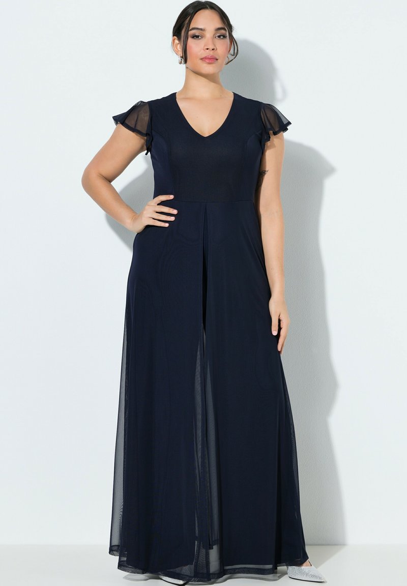 Donna con un abito blu navy lungo fino a terra con maniche trasparenti svolazzanti, in piedi con una mano sul fianco contro uno sfondo bianco.