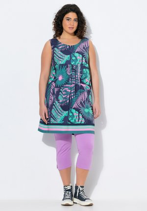 Mouwloze jurk met bloemmotief in marineblauw met groene en roze accenten, gecombineerd met lavendelkleurige capri legging en marineblauwe hoge sneakers.