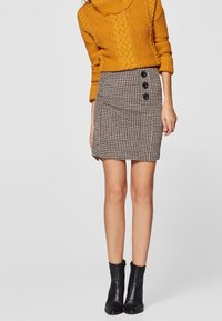 Suéter amarillo de punto con cuello tipo cowl, encima de una falda de cuadros en blanco y negro que presenta tres botones negros en el lado, combinado con botas negras de tobillo.