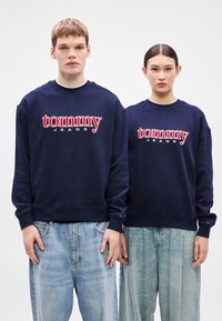 Námornícky modré mikiny s rebrovanou štruktúrou a červenou vyšívanou potlačou "tommy JEANS". Nosené cez svetlomodré široké rifle.