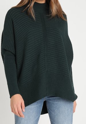 Femme portant un pull en maille côtelée vert foncé à manches longues et col montant, associé à un jean bleu clair.
