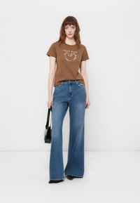 T-shirt en coton marron avec un graphisme décoratif, associé à un jean en denim bleu à jambes larges. Les accessoires comprennent un petit sac à main noir.