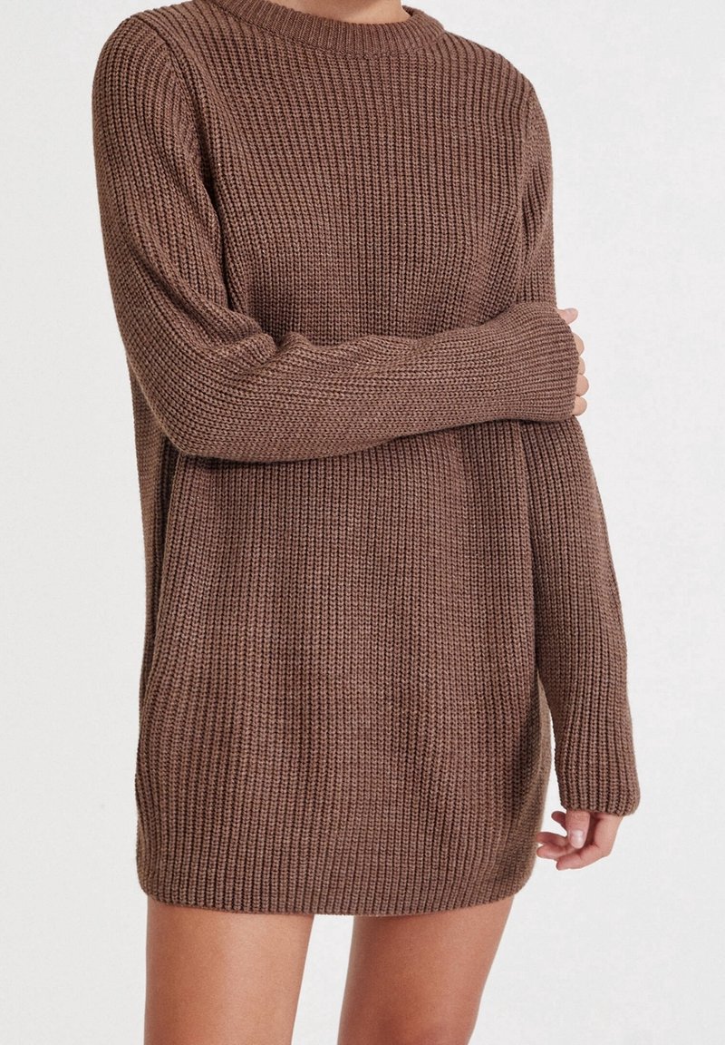 Personne portant une robe-pull marron tricotée, ample, à manches longues, avec un bras croisé et l'autre le long du corps.