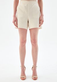 Béžové tweedové šortky s texturovaným vzorem, délka do poloviny stehna a elastický pas. Oblečené s nahými sandály na vysokém podpatku.