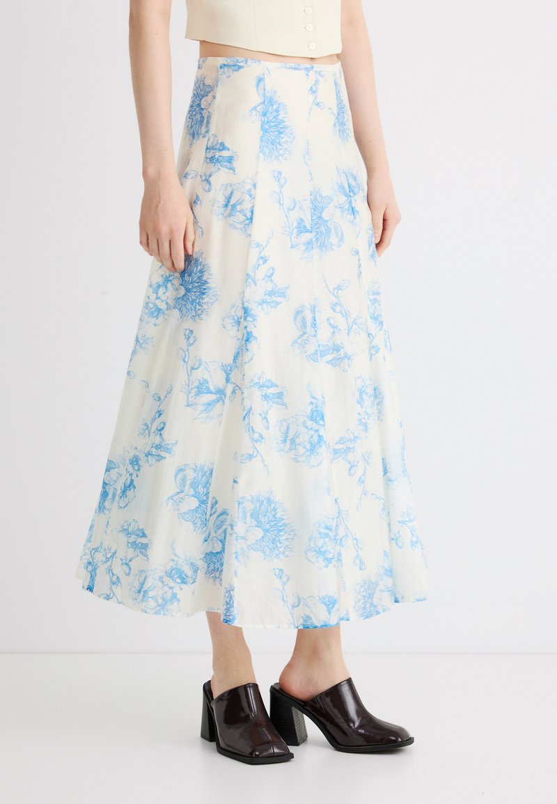 Γυναίκα φοράει λευκή μίντι φούστα με μπλε λουλουδάτο σχέδιο, μπεζ κοντό τοπ και σκούρα καφέ παπούτσια με τακούνι, στέκεται σε λευκό πάτωμα.