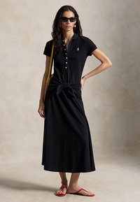 Žena, ktorá má na sebe čierne pološaty s gombíkmi a predným uzlom, čierne slnečné okuliare, žltú kabelku na rameno a červené sandále, pózuje na neutrálnom pozadí.