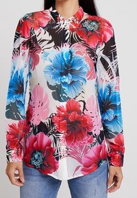 Chemise à boutons avec un motif floral de fleurs bleues et rouges sur un fond blanc translucide. Manches longues, col et ourlet droit.