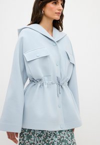 Chaqueta de gran tamaño de color azul claro hecha de un tejido suave, con un gran cuello, dos bolsillos frontales, cierre de botones y una cintura ajustable con cordón.