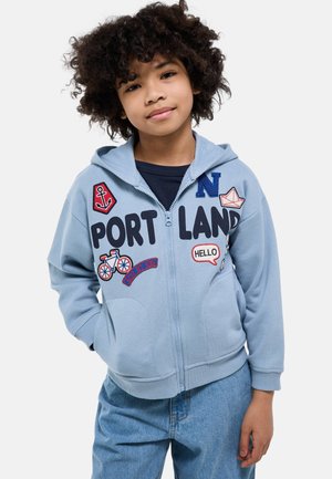 Enfant aux cheveux bouclés portant un sweat à capuche zippé bleu clair avec des écussons indiquant "PORTLAND", une ancre, un vélo et un bateau en papier, associé à un jean bleu.