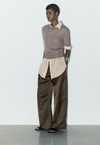 Lekki top w kolorze taupe założony na beżową koszulę z guzikami, w zestawieniu z szerokimi, ciemnobrązowymi spodniami. Gładkie tekstury materiałów; minimalistyczne detale wzornicze.